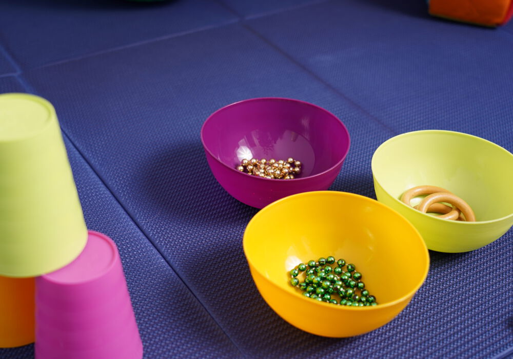 Auf einer blauen Sportmatte stehen im Vordergrund links drei farbige Becher aufgetürmt und recht drei bunte Schüsseln mit Ketten und Ringen