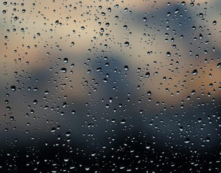 Nahaufnahme von einer Glasscheibe. Auf der Scheibe sind viele Regentropfen. Der Hintergrund ist dunkel und verschwommen.