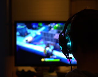 Im Vordergrund ist ein Kopf von hinten zu sehen. Die Person hat Kopfhörer aus. Die Person sitz vor einem Computerbildschirm.