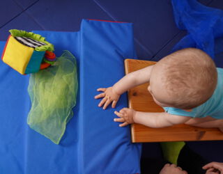 Blick von oben ein Baby, welches eine Holzrampe nach oben krabbelt. Vor dem Kind liegen ein hell-grünes, transparentes Tuch und ein bunter Spielzeugwürfel