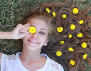 Lächelndes Mädchen liegt auf Gras und hält eine gelbe Blume vor ein Auge, weitere gelbe Blumen liegen in ihrem ausgebreiteten Haar.