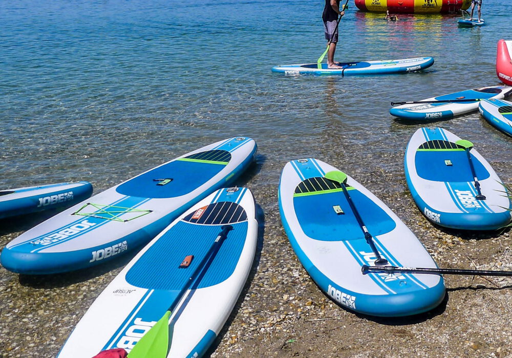 acht blau-weiße Stand Up Paddeling Boards liegen am Ufer eines Sees. Im Hintergrund steht ein Mensch auf einem blau-weißen Board
