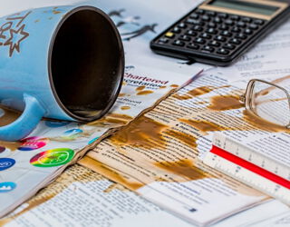 Auf einem Tisch liegen viele Papiere, ein Taschenrechner, ein Lineal, eine Brille und eine umgefallene Kaffetasse. Der Kaffe hat sich über die Papiere verteilt.
