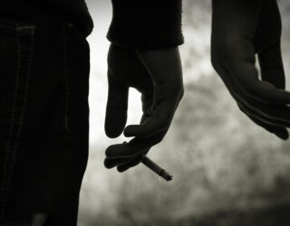 Blick auf zwei Hände von zwei Personen. Die linke Hand hält eine Zigarette. Das bIld hat eine düstere Atmosphäre.