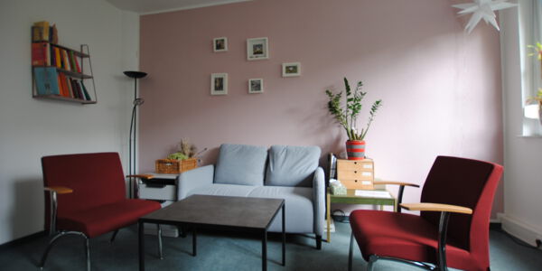 zwei rote Stühle, ein Tisch und ein graues Sofa stehen in einem gemütlich wirkenden Raum mit einer Pflanze und Bildern an den Wänden