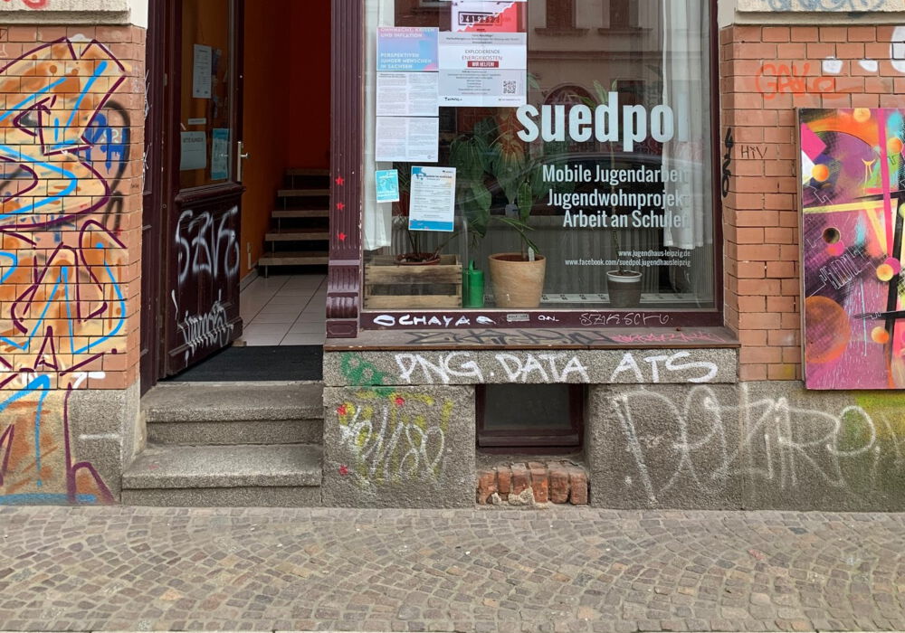 Blick auf die offene Eingangstür des Kontaktladens von suedpol. Im Schaufenster hängen verschiedene Plaktate und Flyer. Rechts und links sind Graffitys an den Hauswänden.