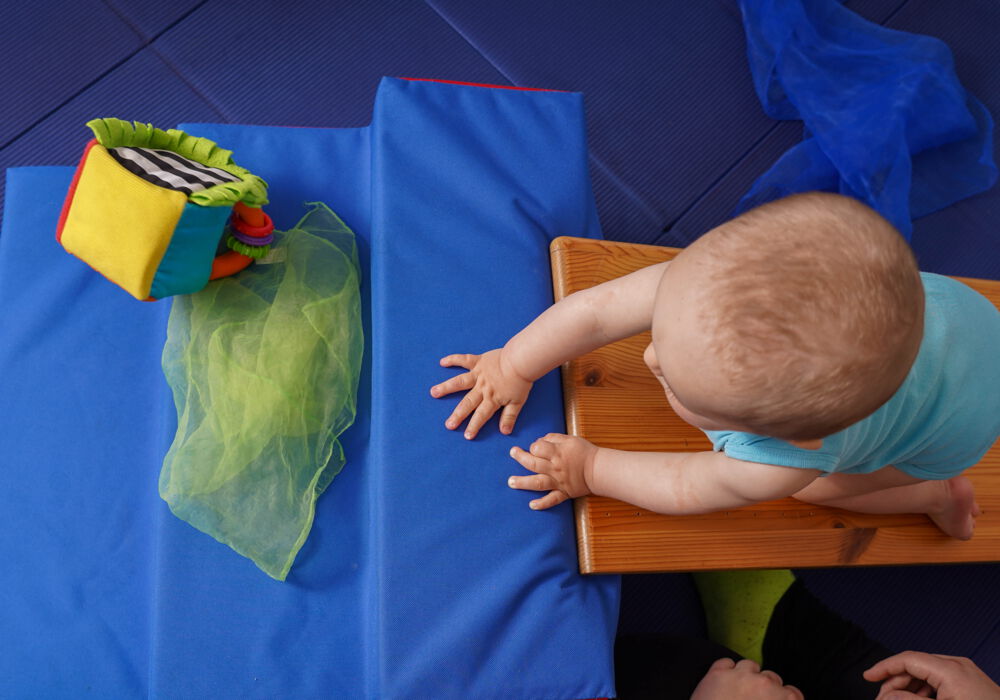 Blick von oben ein Baby, welches eine Holzrampe nach oben krabbelt. Vor dem Kind liegen ein hell-grünes, transparentes Tuch und ein bunter Spielzeugwürfel
