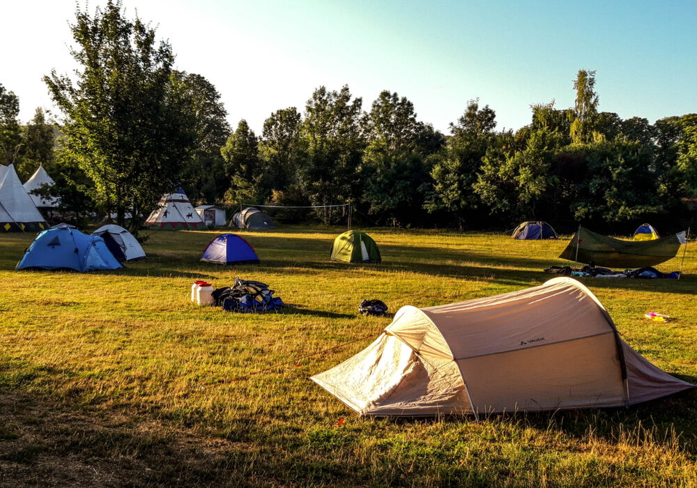 eine weitläufige Wiese im Sommer vor grünen Bäumen, auf der verstreut verschiedene Zelte stehen