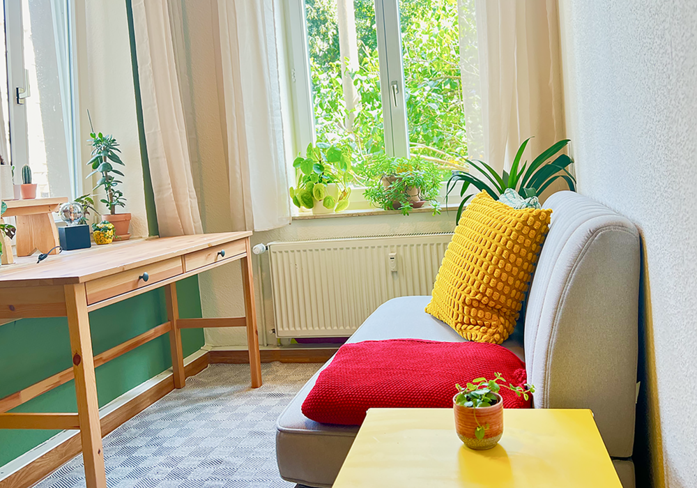 Blick auf eine graue Couch mit einem gelben und roten Kissen und einem gelben Beistelltisch rechts im Bild. Links steht unter einem Fenster ein Holztisch mit Pflanzen.