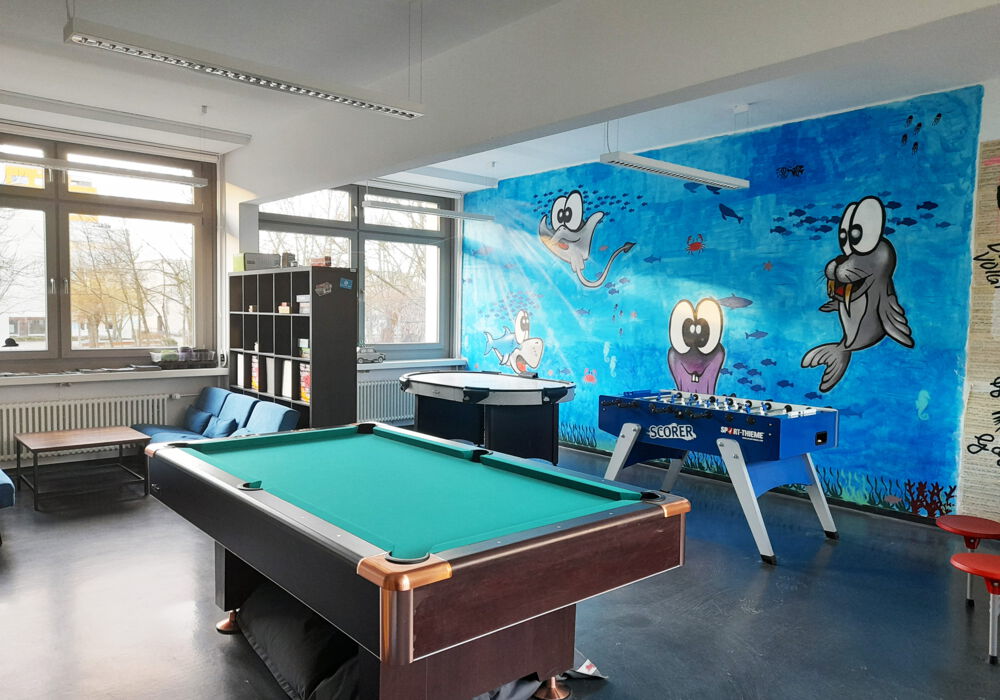 Ein Billiardtisch, ein Kicker und ein Airhockey Tisch stehen vor einer bunten Graffitiwand mit einer Robbe, einem Rochen und einem Hai im Meer