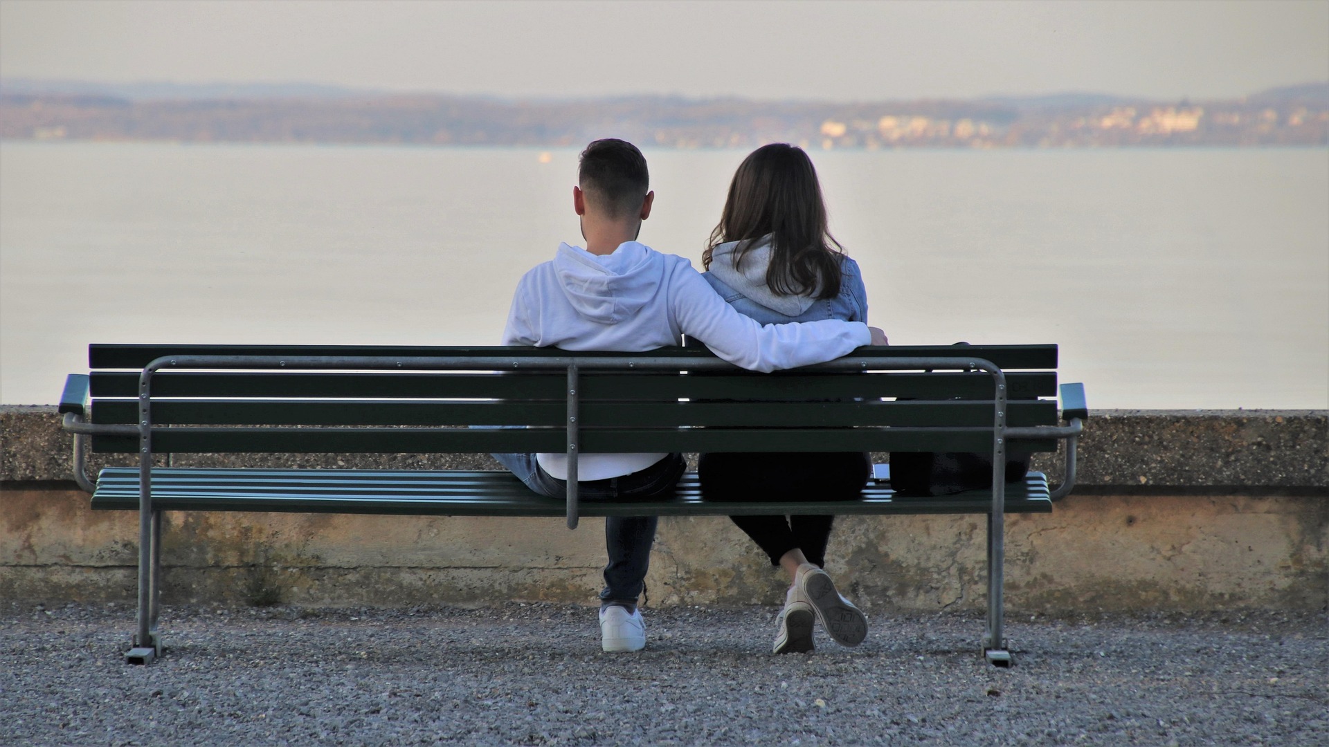 Fotografie zeigt zwei Personen, die auf einer Bank am Ufer sitzen und auf ruhiges Wasser blicken. Szene vermittelt ruhige, intime Atmosphäre mit natürlichem Hintergrund und gedämpften Farben.